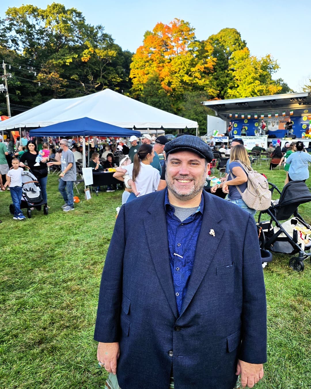 Ryan Michael Kelly sonríe en el Festival Irlandés de Hamilton con asistentes, carpas, un escenario y árboles otoñales detrás de él