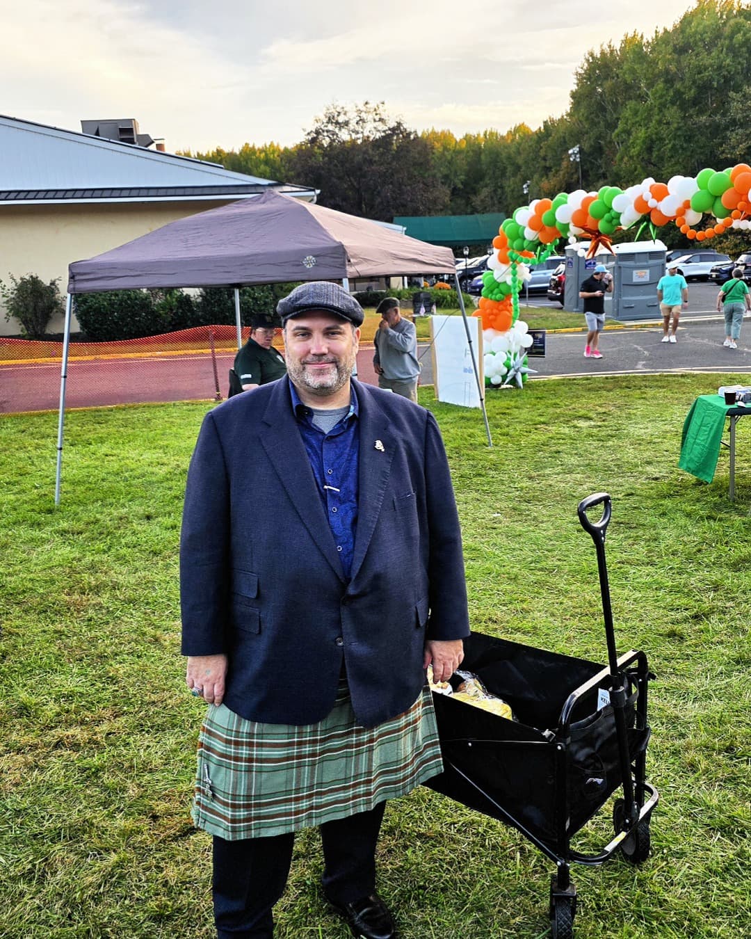 Ryan Michael Kelly con un kilt de tartán verde en el Festival Irlandés de Hamilton cerca de un arco de globos naranja, blanco y verde