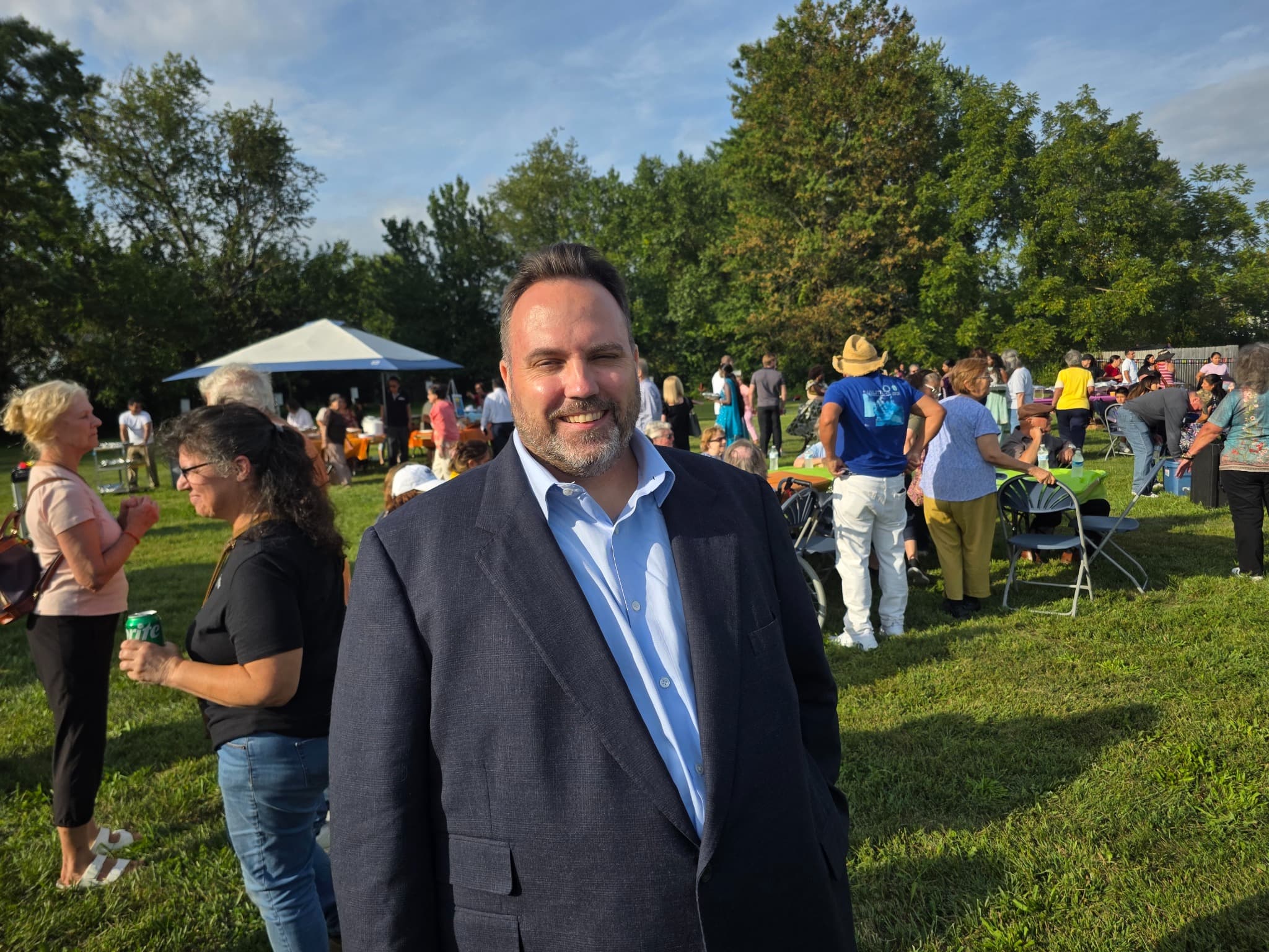 Ryan Michael Kelly smiling outdoors at St. Anthony of Padua’s Multicultural Festival in Hightstown, with community members gathered behind him